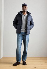 Puffer jacket in navy, gray polo, blue jeans, and brown shoes. Jacket features a hood and zip closure. Model stands on wooden floor.