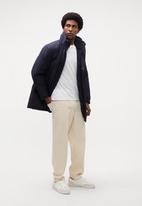 Giacca puffa navy con colletto alto, chiusura a bottoni, indossata sopra una maglietta bianca e pantaloni chino crema, abbinata a sneaker bianche.