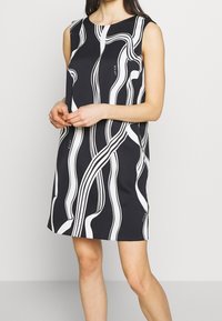 Musta hihaton mekko valkoisilla aaltoilevilla viivoilla. A-linjainen muotoilu, polvimittainen, sileä pinta ja pyöreä pääntie. Ei näkyviä kiinnityksiä.