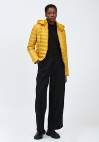 Puffjacke in leuchtendem Gelb mit gestepptem Design und Kapuze, kombiniert mit schwarzen hochgeschnittenen Hosen und schwarzen Ankle Boots.