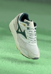 Bijela sportska tenisica s mrežnim i brušenim materijalima, zelenim naglasima, dizajnom na vezivanje i teksturiranom gumenenom potplatu, prikazana na mekoj zelenoj pozadini.