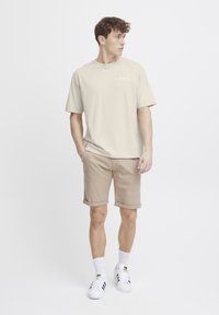 Beige bomulls t-skjorte med grafikk og korte ermer, kombinert med lysebrune shorts med oppbrett. Brukt med hvite sneakers og mønstrede sokker.