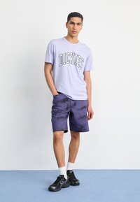 Světle fialové bavlněné tričko se značkovým logem "DICKIES", doplněné tmavě fialovými šortkami s tie-dye vzorem. Černé sportovní boty a bílé ponožky dotvářejí celkový vzhled.
