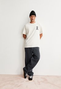 Witte t-shirt met korte mouwen en een donker logo, gecombineerd met donkere, losvallende jeans en zwarte sneakers met witte veters. Zwarte beanie op.