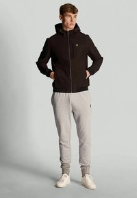 En ung man står med händerna i fickorna iförd en svart huva jacka, ljusgrå sweatpants, stickade strumpor och vita sneakers mot en grå bakgrund.