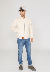 Veste bomber crème clair avec une fermeture éclair, des poignets côtelés et une étiquette orange. Portée avec un jean bleu et des chaussures à lacets marron. Casquette grise avec le logo 'B'.