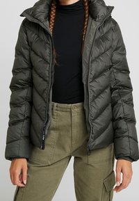 Doudoune vert foncé avec capuche, présentant un motif matelassé diagonal et une fermeture éclair frontale. Associée à un pantalon cargo vert olive.