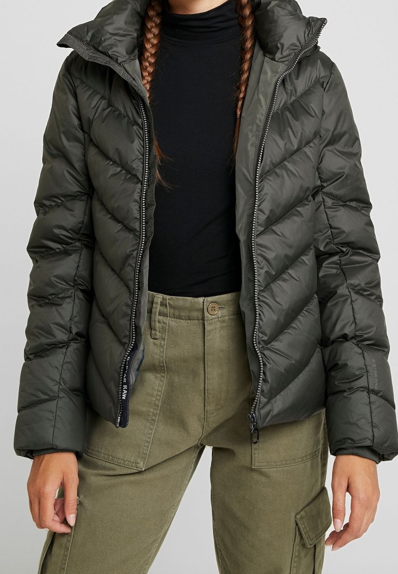 Doudoune vert foncé avec capuche, présentant un motif matelassé diagonal et une fermeture éclair frontale. Associée à un pantalon cargo vert olive.