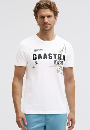 Mann trägt ein weißes kurzärmliges T-Shirt mit schwarzen und orangefarbenen nautischen Grafiken und Texten, kombiniert mit hellblauen Shorts.