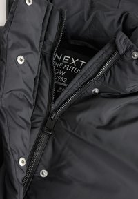 Veste matelassée noire avec fermeture éclair, pressions métalliques et doublure intérieure douce. Étiquette indiquant « NEXT THE FUTURE ». Texture lisse.