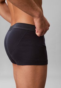 Close-up van een persoon die zwarte Calvin Klein boxers draagt, staand met één hand rustend nabij de tailleband tegen een effen achtergrond.
