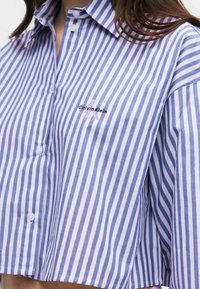 Femme portant une chemise courte Calvin Klein à rayures verticales bleues et blanches, boutonnée avec col.