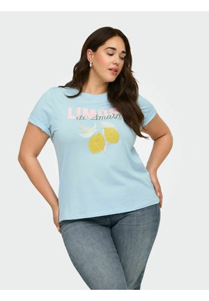 Kobieta o długich ciemnych włosach, ubrana w jasnoniebieski T-shirt z grafiką cytryny i tekstem, w połączeniu z niebieskimi dżinsami, pozująca z rękami na biodrach.