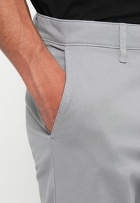 Lichtgrijze broek met een gladde textuur, voorzien van een zijzak en gestikte details langs de tailleband. Verstelbare knoopsluiting.
