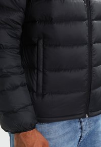Giubbotto imbottito nero con chiusura a zip e tasca laterale, caratterizzato da una texture trapuntata e un tessuto liscio. Jeans blu visibili sotto.