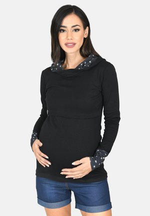 Schwangere Frau in schwarzem Langarmshirt mit Sternen auf den Bündchen und Kapuze, die ihren Bauch hält, trägt Jeansshorts vor hellem Hintergrund.