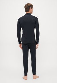 Homme debout pieds nus, tourné de dos, portant un haut ajusté à manches longues noir et un pantalon assorti sur un fond blanc uni.