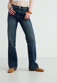 Jeans in denim blu a gamba flare con vita alta, tasche anteriori e chiusura a bottone, abbinati a scarpe in suede color tan. Texture liscia, stile casual.