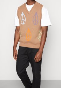 Beiger Strickweste mit V-Ausschnitt, versehen mit bunten Blattmustern in Blau, Lila, Orange und Grün. Getragen über einem weißen T-Shirt.