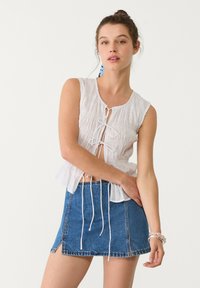 Weißes ärmelloses Top mit Frontknüpfung, strukturierter Stoff; kombiniert mit einem kurzen Jeansrock mit fransigem Saum und seitlichen Schlitzen.