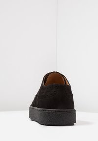 Černé semišové boty s detaily v brogue stylu, texturovanou gumovou podrážkou a prošitými akcenty. Zadní pohled ukazuje hladké podšívání a klasický design.