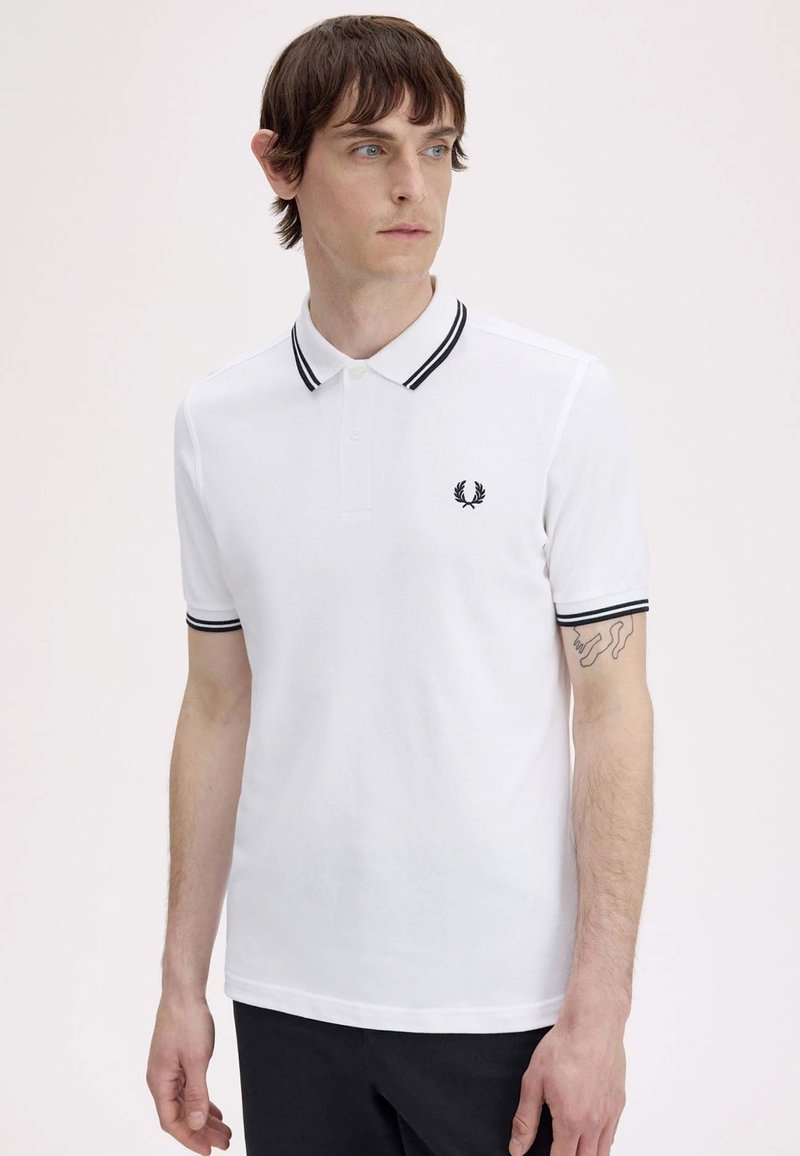 Jeune homme aux cheveux bruns courts portant un polo blanc avec des bordures noires et un petit logo laurier noir sur la poitrine, debout devant un fond uni.