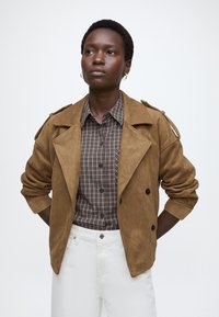 Personne portant une veste en daim marron sur une chemise à carreaux et un pantalon blanc, debout les mains derrière le dos sur un fond uni.