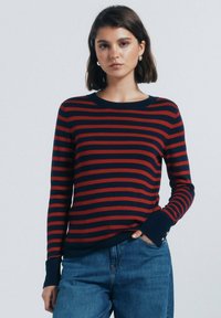 Rood en marineblauw gestreepte gebreide trui met een ronde halslijn, ribbelmanchetten en knoopaccenten op de mouwen. Draagt met denim jeans.
