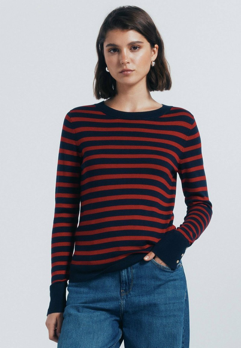 Rood en marineblauw gestreepte gebreide trui met een ronde halslijn, ribbelmanchetten en knoopaccenten op de mouwen. Draagt met denim jeans.