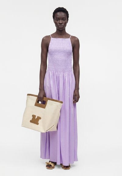 Γυναίκα που φοράει ένα μίντι λιλά φόρεμα χωρίς μανίκια και σανδάλια, κρατώντας μια μεγάλη μπεζ τσάντα tote με καφέ λαβές και λογότυπο.