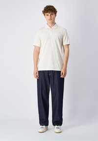 Cremfarbene Polohemd mit Dreiknopfleiste, kombiniert mit lässigen dunkelblauen Jeans. Weiße Sneaker runden das Outfit ab.
