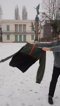 Άτομο που πετά ή περιστρέφει ένα πράσινο και μαύρο μπουφάν σε εξωτερικό χώρο σε χιονισμένο έδαφος κοντά σε άγαλμα και κτίριο με κλειστά παραθυρόφυλλα.