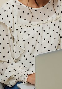 Weißes Bluse mit Punkten, Rüschensaum und gerüschten Ärmeln, schwarz gepunktet und mit zarten Rüschen am Halsauschnitt.