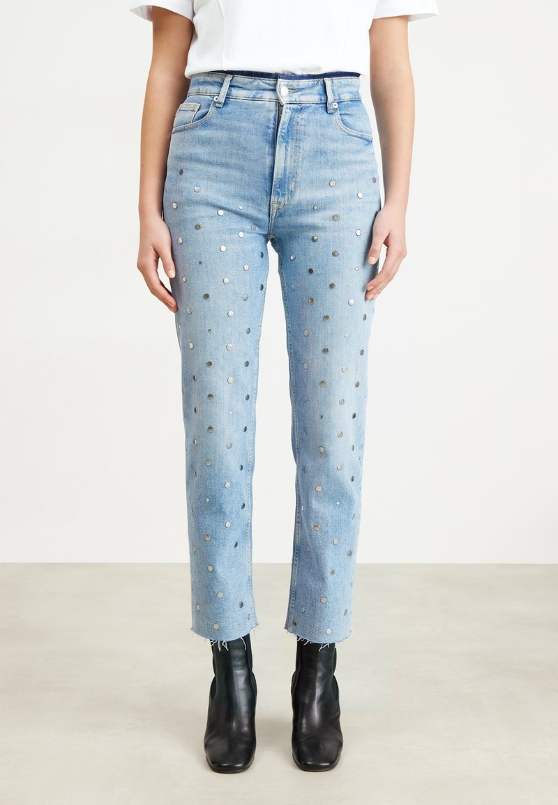 Jean bleu clair taille haute avec des clous circulaires argentés partout, ourlets effilochés, porté avec des bottines noires et un haut blanc à manches courtes.