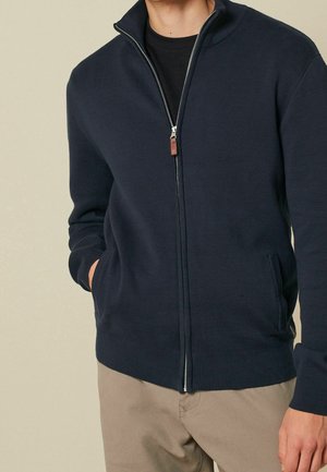 Homme portant un pull zippé bleu marine avec poches par-dessus une chemise noire et un pantalon beige, mains dans les poches, fond neutre.