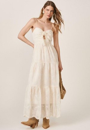 Femeie într-o rochie maxi albă fără mâneci cu broderie florală, ținând o geantă bej, purtând botine bej și cercei deosebiți.