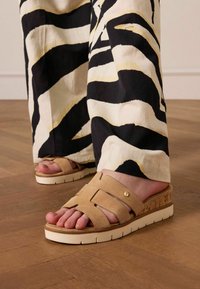 Lichtbruine gamuza sandalen met een kurken wedge zool en witte rubberen buitenzool, voorzien van kruislingse banden en een gouden accentdetail.