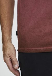Burgunderfarget T-skjorte med et mykt, teksturert stoff, som har en rett passform og synlig kant. Hånd plassert nær kanten av skjorten.