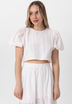 Bílý bavlněný crop top s nafouklými rukávy, zdobený detaily z krajky a vertikálními vzory. Doplněn ladící krajkovou sukní.