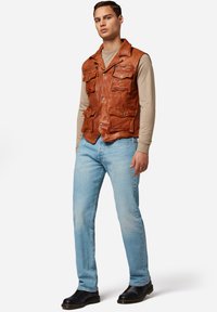 Braune Lederweste mit mehreren vorderen Taschen, Knopfverschluss und Kragen-Design, getragen über einem beigen Langarmshirt und blauen Jeans.