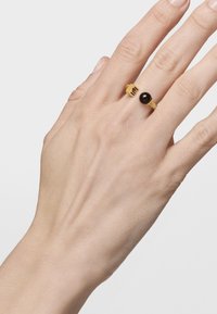 Anillo de oro con una piedra negra redonda, banda texturizada y diseño decorativo parecido a una corona en los lados. Llevado en una mano delgada.