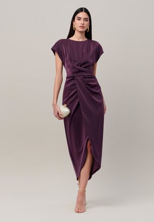 Vrouw draagt een paarse satijnen midi-jurk met geplooide voorkant, houdt een kleine ronde clutch vast, draagt gouden oorringen en nudekleurige sandalen.