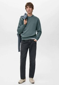 Felpa verde con scollo tondo, jeans neri e sneakers chiare. Una giacca blu è appoggiata a un braccio. Sfondo semplice.