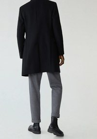 Langer schwarzer Mantel aus strukturiertem Stoff, kombiniert mit grau gekrempelten Hosen und schwarzen Stiefeletten. Besitzt eine taillierte Passform und ein minimalistisches Design.