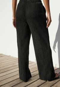 Pantaloni negri cu un croi larg, dintr-un material texturat, având un brâu elastic. Văzuți din spate, arătând o croială lejeră.