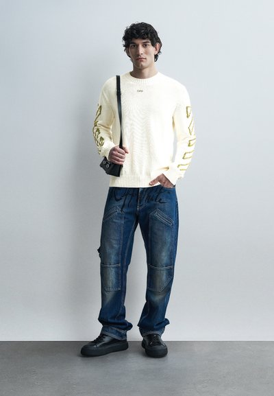 Άνδρας με κατσαρά μαλλιά, φοράει εκρού πουλόβερ με χρυσά γράμματα, μπλε τζιν με μαύρη γραφή, μαύρα παπούτσια και μια μικρή μαύρη τσάντα ώμου.