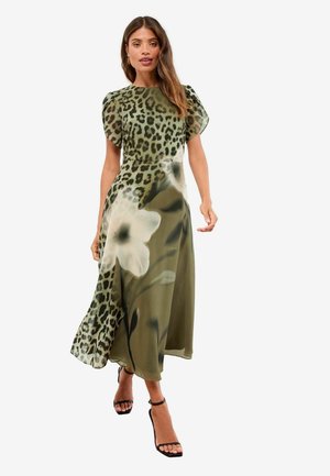 Žena v zelenej midi šaty so zmiešaným leopardím vzorom a veľkým bielym kvetinovým vzorom, krátkymi nafúknutými rukávmi a čiernymi sandálmi na vysokom opätku.
