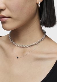 Collane a strati con un collier in argento spesso e una delicata catena in argento con un piccolo pendente nero. Finitura metallica liscia.