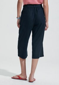 Jūros mėlynumo trumpi keliaraiščiai su laisvu kirpimu, elastinga juosmeniu ir dviem užpakalinėmis kišenėmis. Nešiojami su raudonais plokščiais sandalais, matomos apatinės kojos dalys.