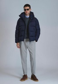 Marineblaues Pufferjackett mit hohem Kragen, zwei Vorderstäbchen, getragen über einem dunklen Pullover und einem hellen Hemd mit Kragen. Graue Hosen und braune Schuhe.